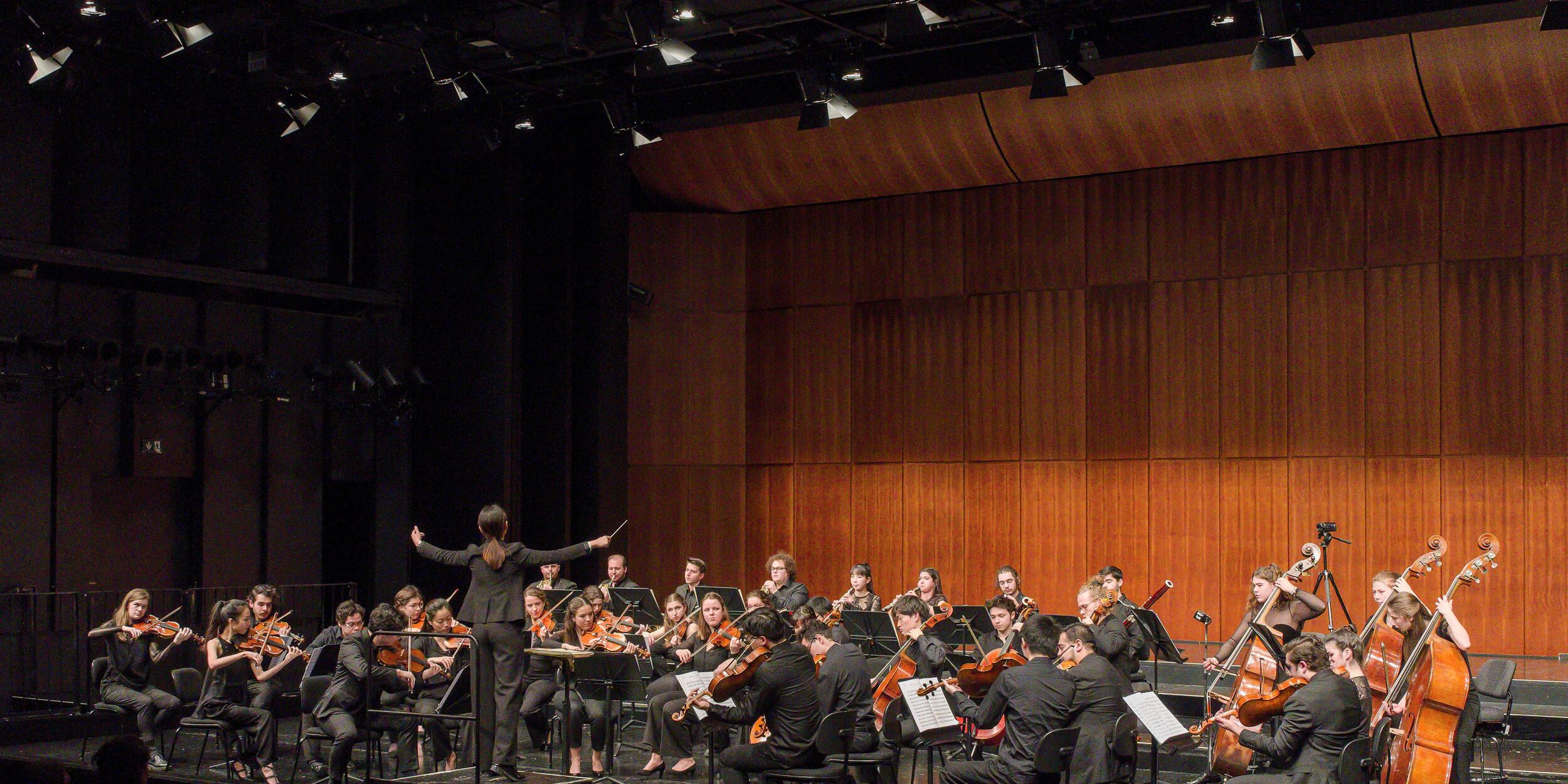 Orchester auf einer Bühne, holzvertäfelte Wand, Dirigentin mit Rücken zum Publikum | © Christian Schneider