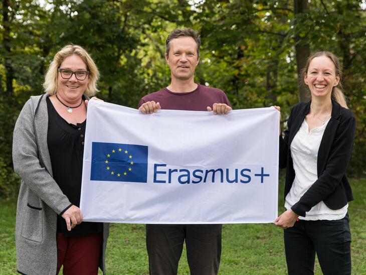 Drei Menschen auf einer Wiese, dahinter Wald, sie halten eine Erasmus-Flagge