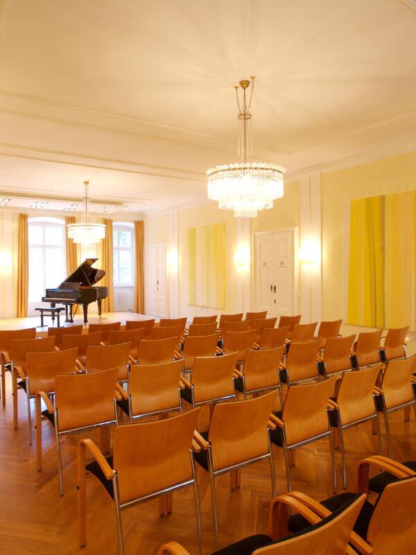 Kleiner Konzertsaal mit Bestuhlung und einem Klavier, gelbliches Licht, Parkettboden