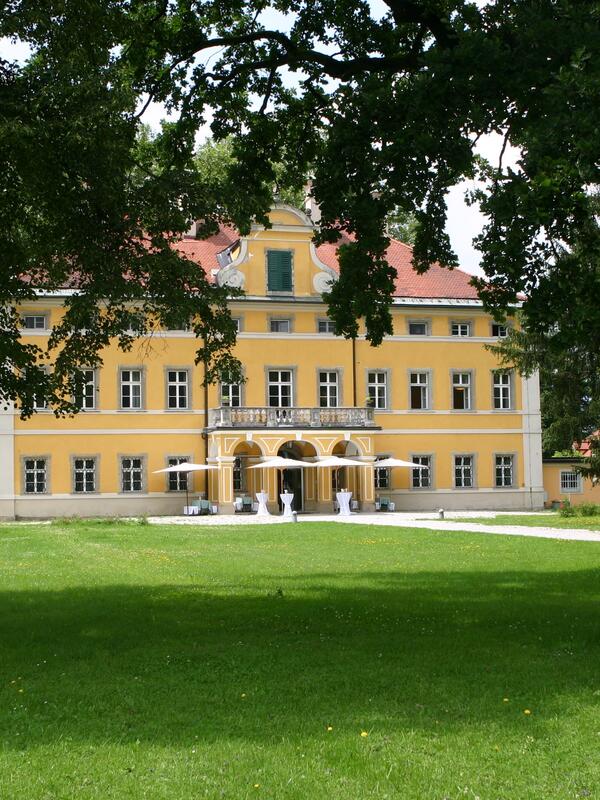 Blick auf die Frohnburg, ein gelbes Schloss in einem Park, an zwei Bäumen vorbei fotografiert