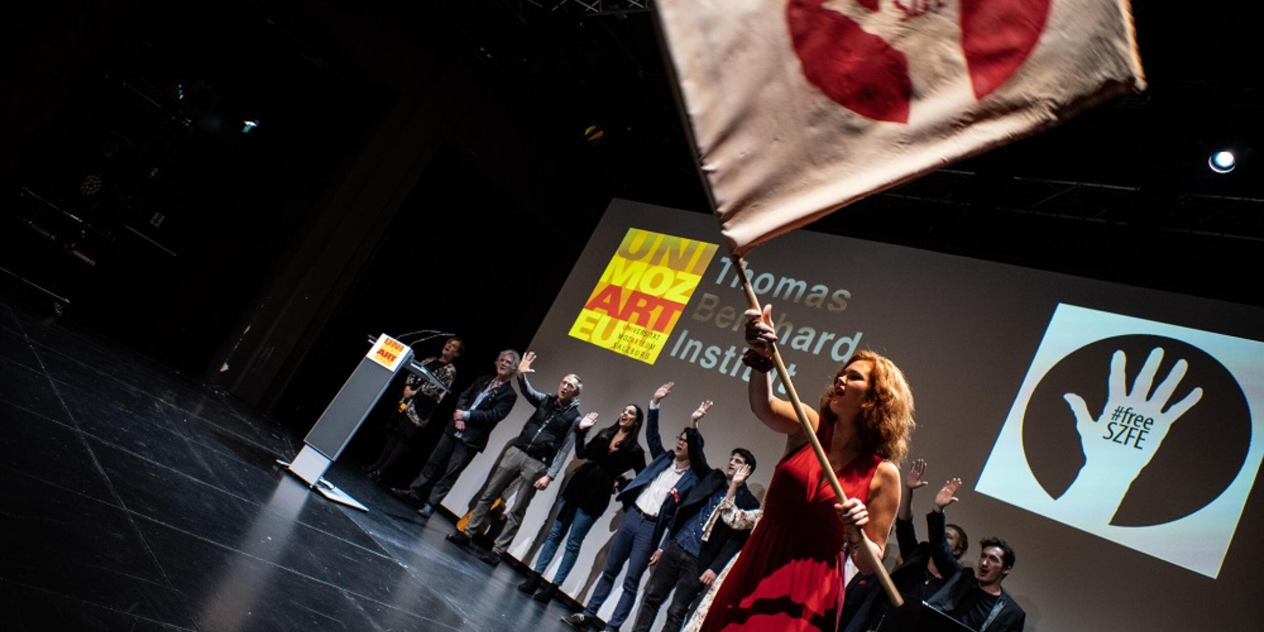 Menschen auf der Bühne, eine Frau in rotem Kleid schwingt eine Fahne mit Aufschrift "Free SZFE" | © Fabian Schober