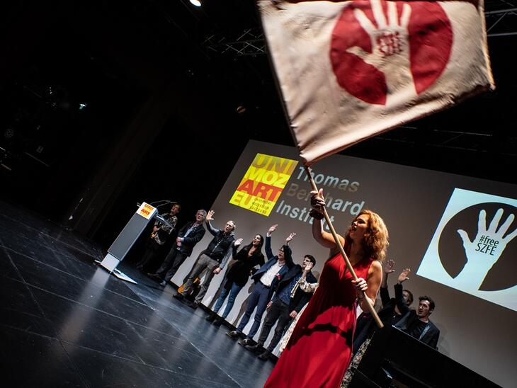 Menschen auf der Bühne, eine Frau in rotem Kleid schwingt eine Fahne mit Aufschrift "Free SZFE" | © Fabian Schober
