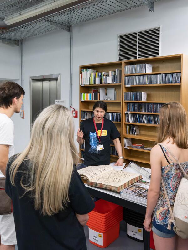 © Christian Schneider Mann vor einem Bücher- und CD-Regal, erklärt für Besucher*innen ein altes großes Buch auf | © Christian Schneider