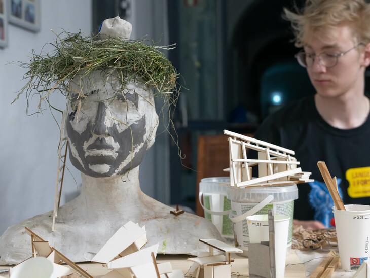 Ein Kopf aus Gips mit Heu auf dem Kopf im Vordergrund, dahinter ein junger Mann und Utensilien zum Modellbau | © Christian Schneider
