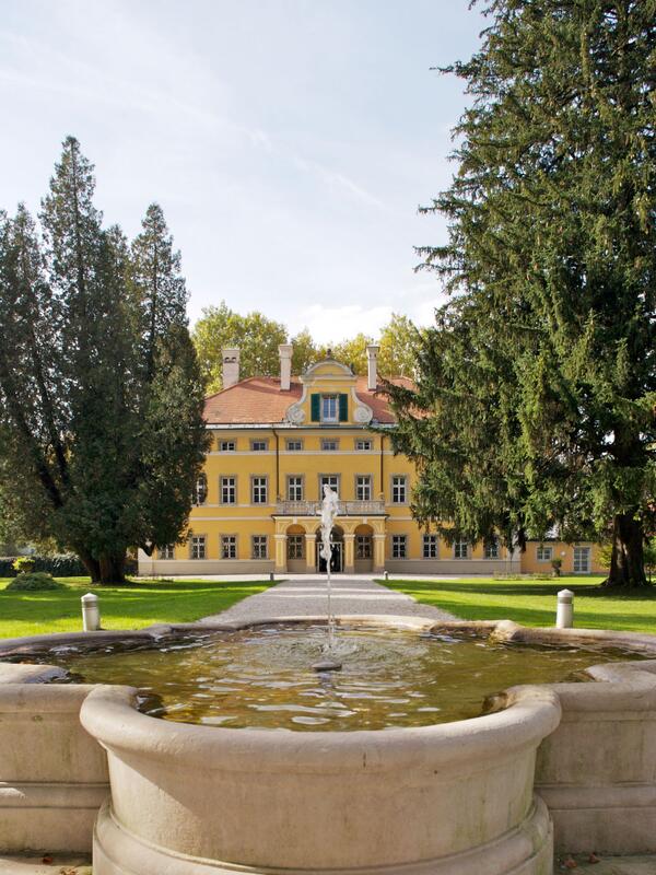 Blick auf die Frohnburg, ein kleines gelbes Schloss in einer sonnigen Parkanlage. Kieswege, Bäume, ein kleiner Brunnen