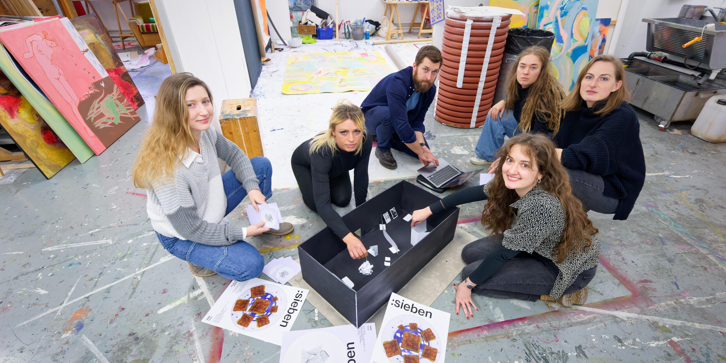 Sechs Studierende in einem Arbeitsraum, Staffeleien, Malereien, am Boden Plakate mit der Aufschrift "sieben" | © Christian Schneider