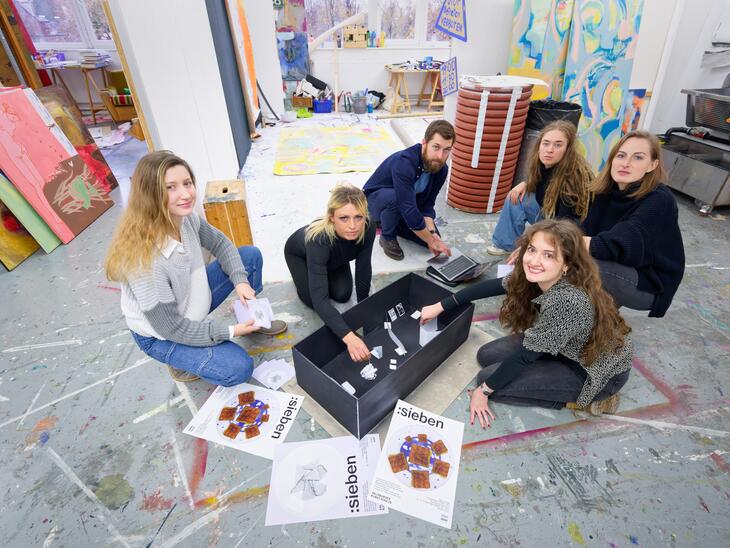 Sechs Studierende in einem Arbeitsraum, Staffeleien, Malereien, am Boden Plakate mit der Aufschrift "sieben" | © Christian Schneider