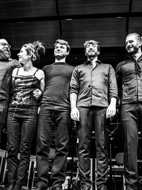 Bild schwarz-weiß, Musiker*innen in einer Reihe auf der Bühne, lachen ins Publikum | © Fabian Schober