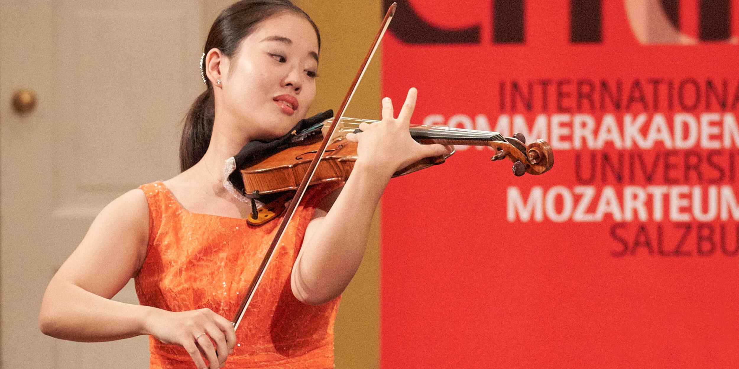 Asiatische Frau in orangem Kleid spielt Violine, roter Hintergrund | © Christian Schneider